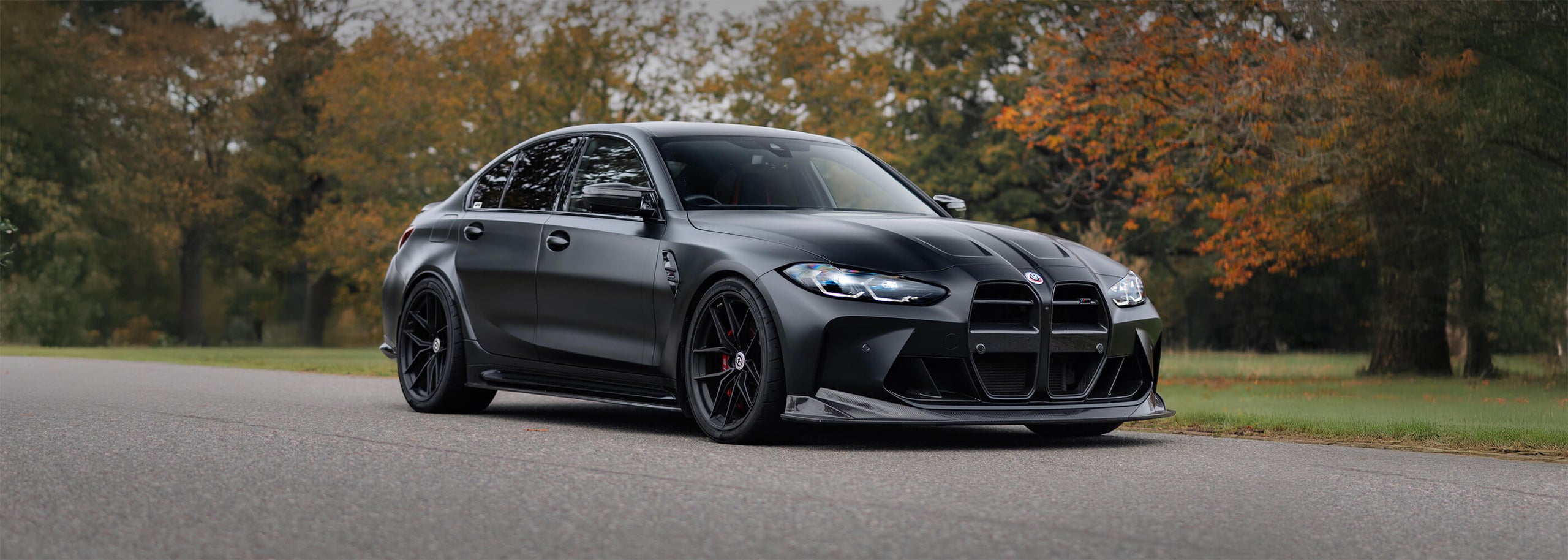 A dark gray BMW M3 sedan with black wheels and a front splitter, photographed on an asphalt road with trees in the background.